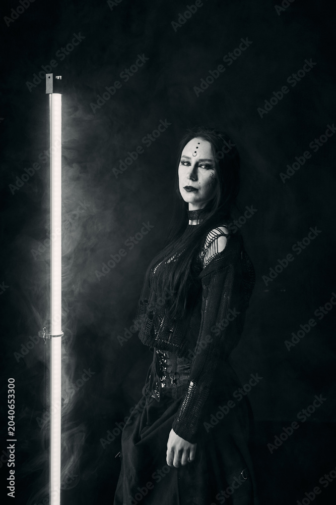 Obraz premium Gothic young woman with long dark hair and black makeup posing by vertical light tube in smoke