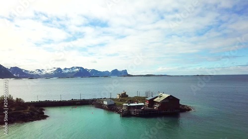 A small village sits by the water. Houses and trailers line the coast. Green hills rise in the background under a clear sky. The calm water reflects the scenery.
