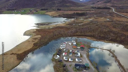 A small village sits by the water. Houses and trailers line the coast. Green hills rise in the background under a clear sky. The calm water reflects the scenery.