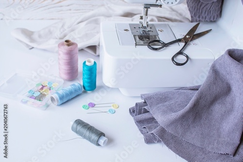 Sewing supplies on a white table with a sewing machine in the background. Sewing machine with fabric and sewing accessories on a white table.