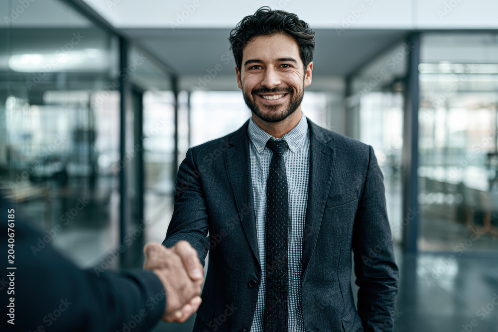 Fototapeta premium A smiling businessman in a suit shaking hands with another person inside a modern office with glass walls and bright lighting.