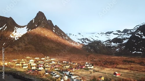 A small town is visible along the coast of Norway. Mountains rise in the background. Sunlight casts a warm glow on buildings. The sky remains mostly clear as the day ends.