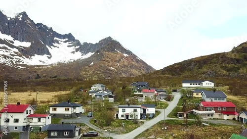 A small town is visible along the coast of Norway. Mountains rise in the background. Sunlight casts a warm glow on buildings. The sky remains mostly clear as the day ends.