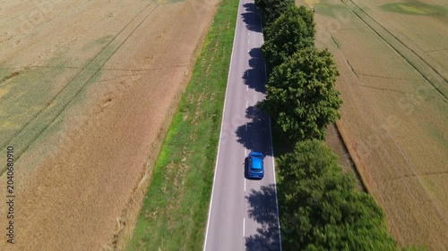 Aerial video of a car driving along a scenic rural road between farm fields 
