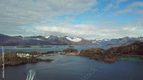 A wide view shows islands surrounded by water with mountains and trees in the background. The sun is shining and the sky is mostly clear. This area is located in Norway.