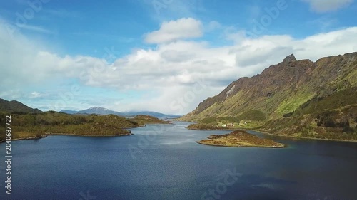 A wide view shows islands surrounded by water with mountains and trees in the background. The sun is shining and the sky is mostly clear. This area is located in Norway.