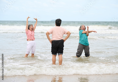 Elderly women exercising on the beach, with gentle waves lapping at their feet. The two women on the sides are stretching