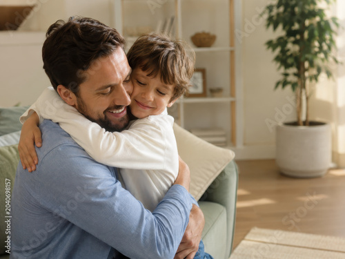Happy father son hug joyful family bond in cozy living room, warm sunlight, casual home clothing, tender embrace, parenting love moment