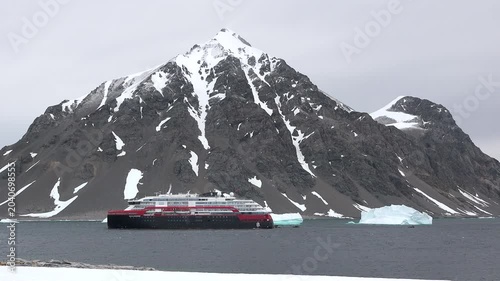 A large cruise ship floats in the cold waters of Antarctica surrounded by snow-covered mountains under gray clouds. Small icebergs are visible in the distance.