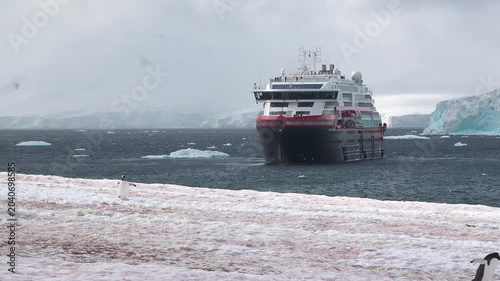 A large cruise ship floats in the cold waters of Antarctica surrounded by snow-covered mountains under gray clouds. Small icebergs are visible in the distance.