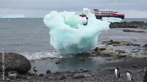 A large cruise ship floats in the cold waters of Antarctica surrounded by snow-covered mountains under gray clouds. Small icebergs are visible in the distance.