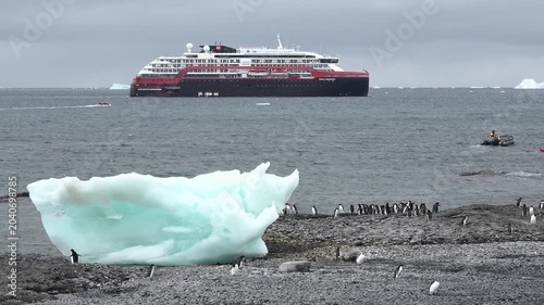 A large cruise ship floats in the cold waters of Antarctica surrounded by snow-covered mountains under gray clouds. Small icebergs are visible in the distance.