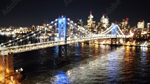 Blurred night view of a suspension bridge spanning across a river with illuminated city skyline skyscrapers bokeh lights, concept for urban travel, metropolis nightlife and cinematic backgrounds
