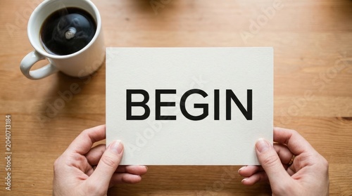 Overhead view of hands holding a card with the word 'begin' next to a steaming cup of black coffee on a wooden surface