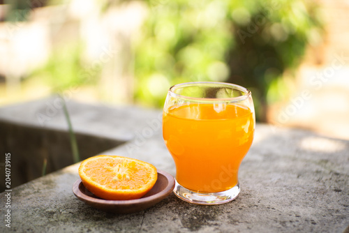 Fresh orange juice in a glass with halved and whole oranges on a stone ledge, bright outdoor bokeh background