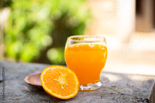 Fresh orange juice in a glass with halved and whole oranges on a stone ledge, bright outdoor bokeh background