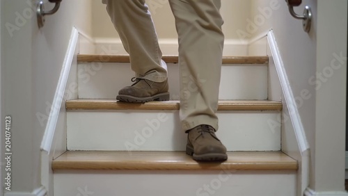 Individual wearing beige pants and brown shoes descends wooden stairs, showcasing foot placement and movement in a well-lit indoor setting with handrails visible