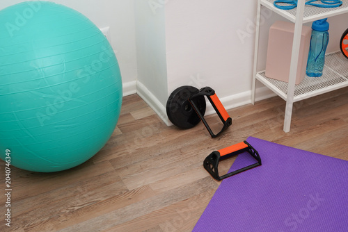 A home gym corner with various fitness equipment. A turquoise stability ball, purple yoga mat, and push-up bars on a wooden floor. Concept of exercise, health, and active lifestyle at home.