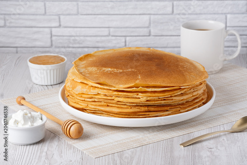 Stack of pancakes served with honey and cream on a table with a cup in a bright kitchen setting