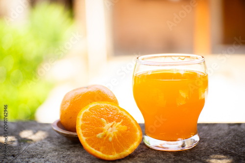 Fresh orange juice in a glass with halved and whole oranges on a stone ledge, bright outdoor bokeh background