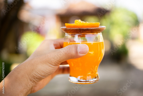 Hand holding glass of fresh orange juice with wooden lid and orange slice outdoors, sunny blurred background