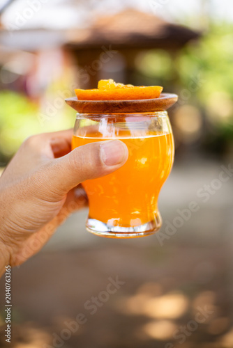 Hand holding glass of fresh orange juice with wooden lid and orange slice outdoors, sunny blurred background