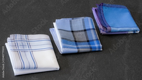 Three different folded men's cotton handkerchiefs in varying blue, white, and plaid patterns  on a textured dark background.