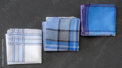 Three different folded men's cotton handkerchiefs in varying blue, white, and plaid patterns  on a textured dark background.