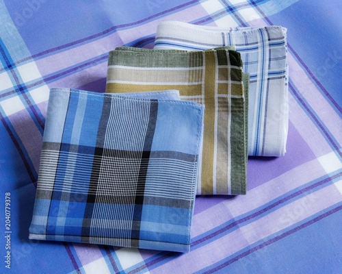 A set of three neatly folded cotton handkerchiefs in varying plaid and checkered patterns is arranged on a blue and purple striped cotton handkerchief.