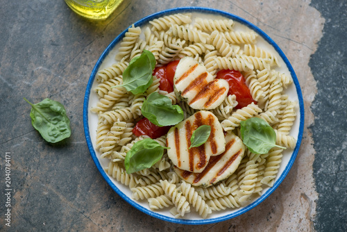 Pasta salad with grilled halloumi and tomatoes on a white and blue plate, horizontal shot on a beige and grey granite background, high angle view