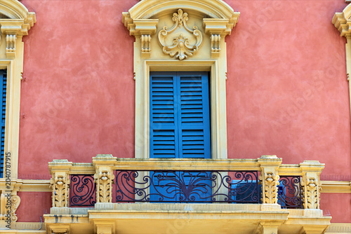 A building with a pink exterior features blue shutters and a decorative balcony. This structure is located in Modena, Italy. Sunlight illuminates the details of the architecture.