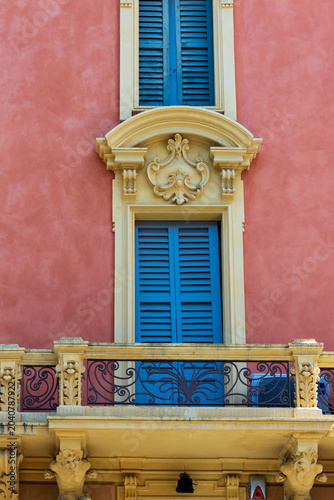 A building with a pink exterior features blue shutters and a decorative balcony. This structure is located in Modena, Italy. Sunlight illuminates the details of the architecture.