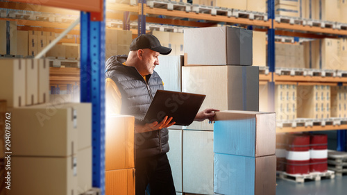 Photos Warehouse logistics, Worker checks cardboard boxes with laptop among storage racks during inventory and order management