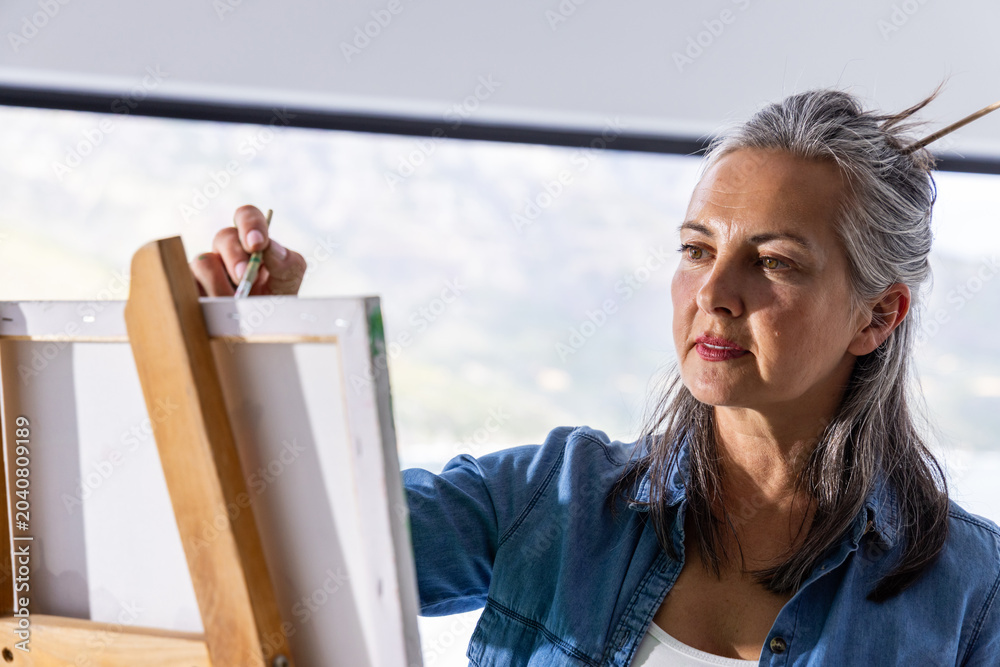 Naklejka premium Senior woman painting on canvas at studio with wooden easel holding brush wearing denim, copy space