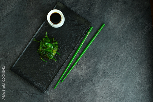 Chuka wakame seaweed salad on a black slate board with a white cup of soy sauce and green chopsticks, healthy Japanese appetizer