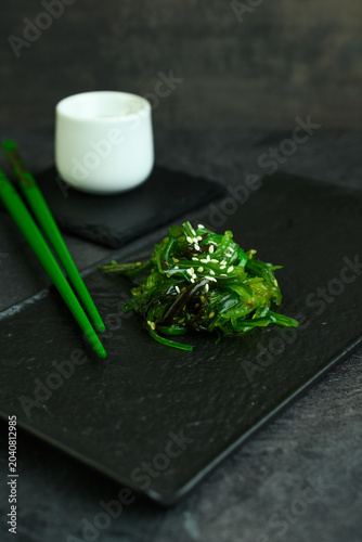 Chuka wakame seaweed salad with sesame seeds on a black slate board with a white cup of soy sauce and green chopsticks, Japanese appetizer