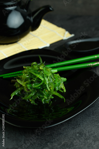 Japanese chuka wakame seaweed salad on a black plate with green chopsticks, black teapot and bamboo mat, traditional Asian table setting