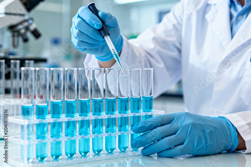 Scientist in lab coat and gloves using pipette to add blue liquid to test tubes in laboratory