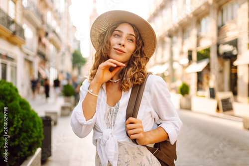 A smiling tourist with a backpack and hat enjoys a sunset stroll. The woman poses on a city street in the sun's rays. Concept of tourism, recreation, and beauty.