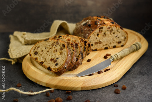 Sliced homemade raisin walnut bread on a round wooden board with a vintage knife, burlap cloth and scattered raisins on a dark rustic background