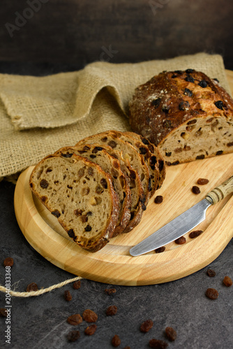 Sliced homemade raisin walnut bread on a round wooden board with a vintage knife, burlap cloth and scattered raisins on a dark rustic background