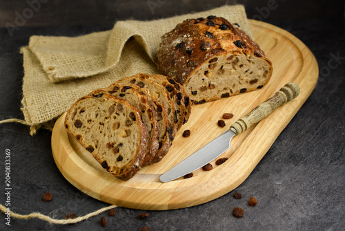 Sliced homemade raisin walnut bread on a round wooden board with a vintage knife, burlap cloth and scattered raisins on a dark rustic background