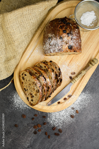 Top view of sliced raisin walnut bread on a wooden tray with a sugar sieve, knife and burlap, flat lay of homemade artisan bakery