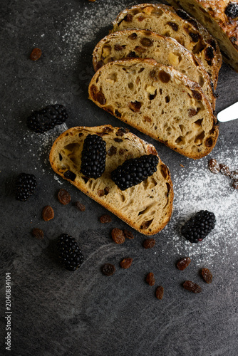Top view of raisin walnut bread slices with fresh blackberries, scattered raisins and powdered sugar on a dark stone, flat lay bakery