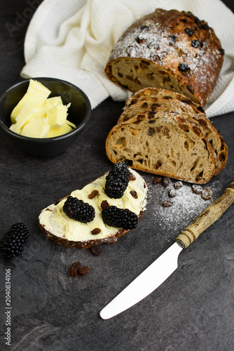 Raisin walnut bread served with butter and fresh blackberries, a bowl of butter and vintage knife on a dark background, rustic breakfast