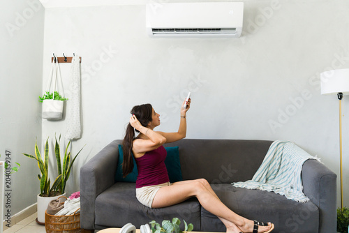 Overheated woman seated on a couch, fixing her hair as she turns on the air conditioning using a remote control