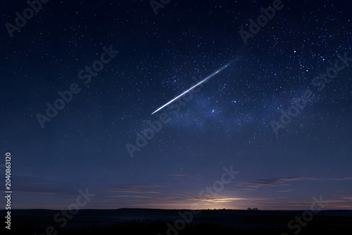 A bright meteor streaks across a star-filled deep blue night sky over a dark landscape, with a soft glow on the horizon.