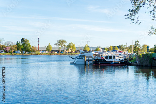 Tableau sur toile Motor yachts and cabin cruisers moored along a calm river canal with blue water reflections, spring trees, harbor infrastructure, and peaceful marina atmosphere under a clear sky