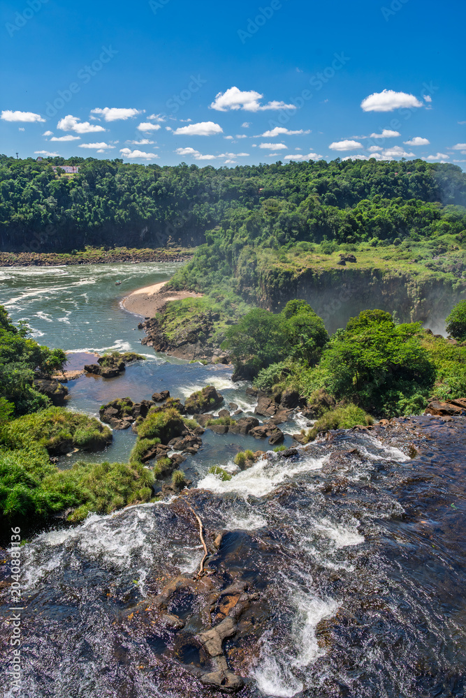 Fototapeta premium Panoramic view of the majestic Iguazu Falls in Iguazu National Park, surrounded by lush tropical rainforest under a clear blue sky with white clouds