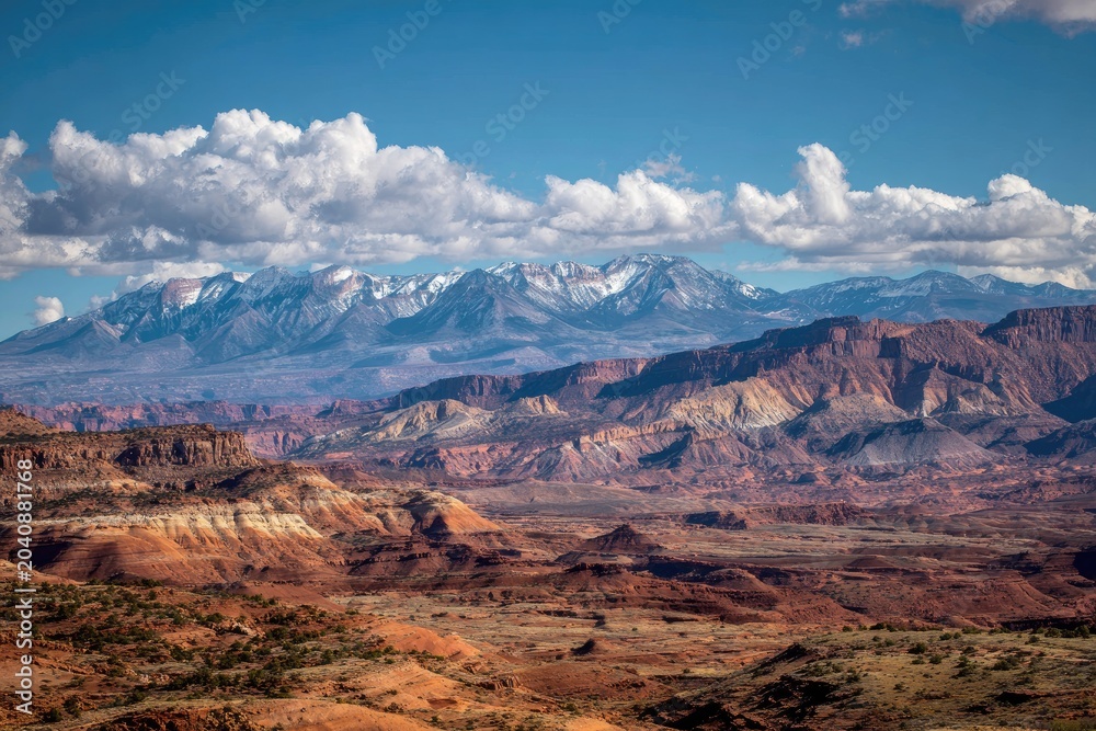 Fototapeta premium Red rock formations under snow-capped mountains, vast desert landscape, cloudy sky
